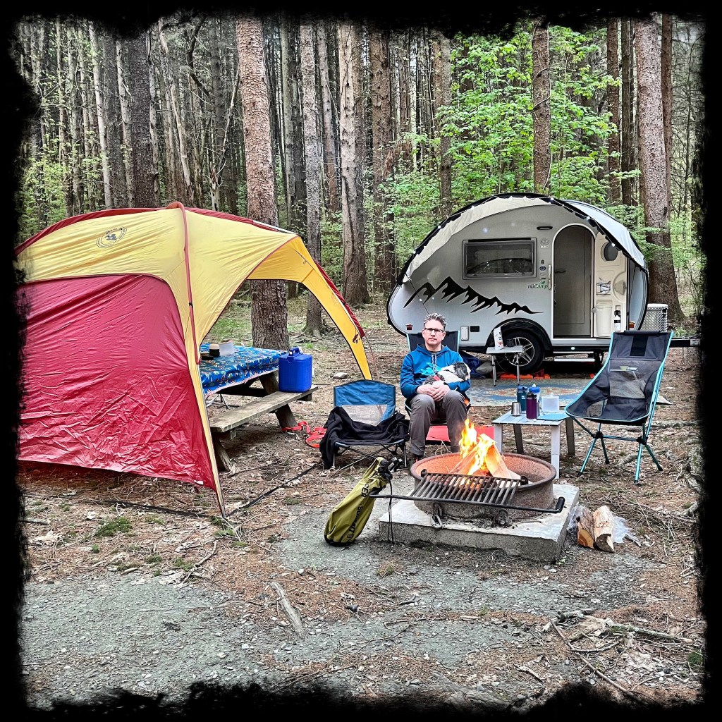 Man and dog sitting in camp chairs by a fire in front of a camper