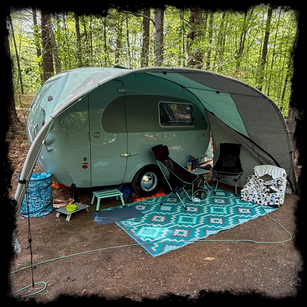 Photograph of our Barefoot Caravan at a wooded campsite, with an awning and three camp chairs sitting on a rug in front of it. 