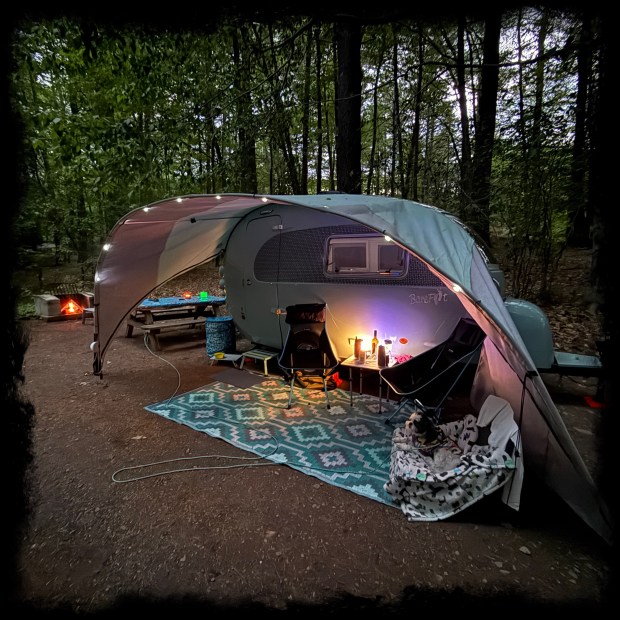 Photograph of the Barefoot outside at night with a fire i the background and lights along the edge of the awning. 